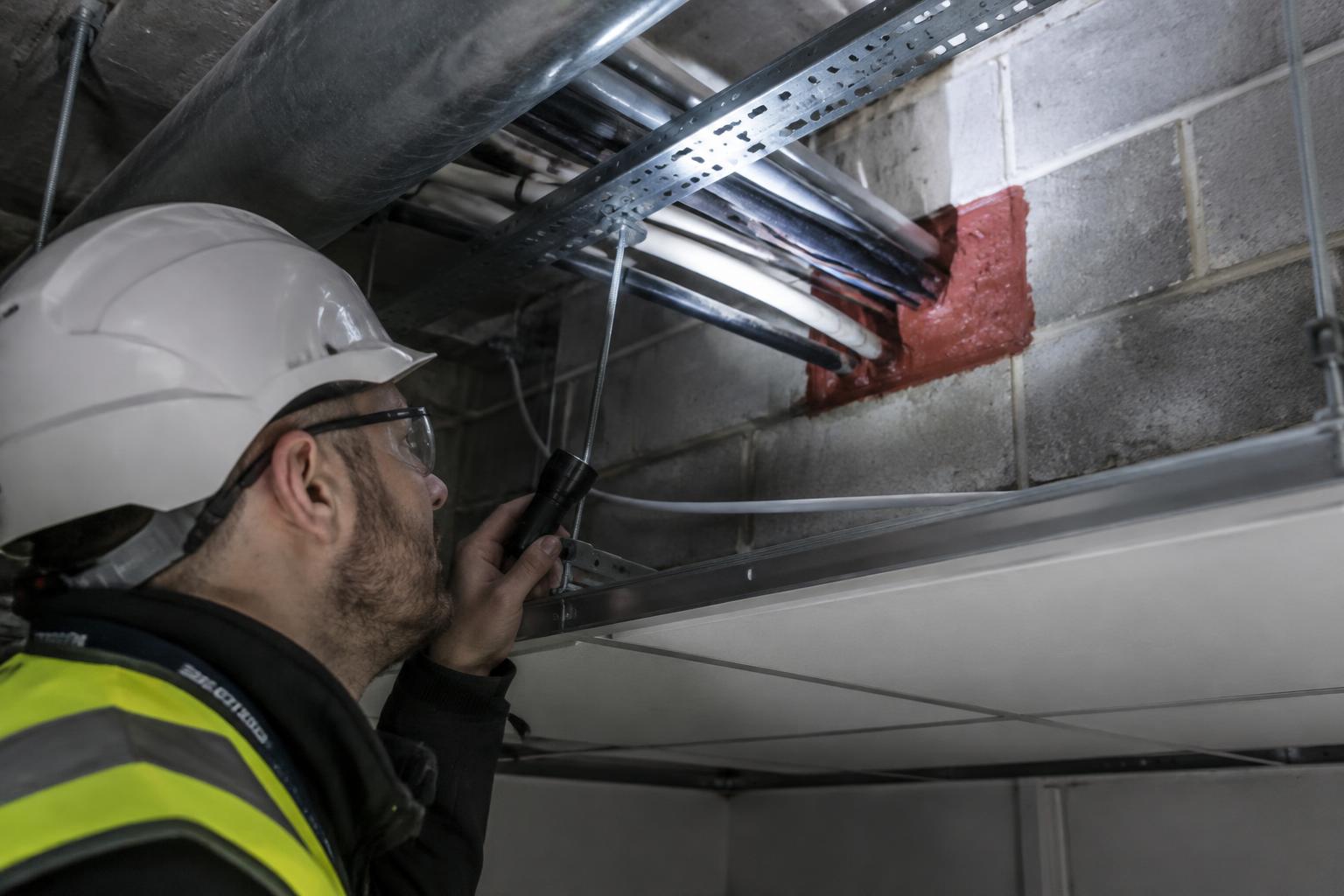 Surveyor with a torch inspecting firestopped cable penetrations in a ceiling void